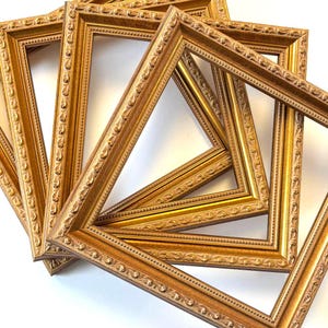 May include: A collection of ornate, golden picture frames. The frames have a detailed, embossed design along the edges and corners. The frames are empty, with a light-colored background visible through the openings. The frames are arranged in a slightly overlapping pattern.