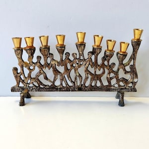 May include: A nine-branch menorah crafted from dark, textured metal featuring a row of stylized human figures linked together. Each branch is topped with a polished, smooth gold-toned candle cup. The base is supported by two feet.