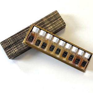 May include: A set of small perfume bottles in a gold-colored tray, with a matching patterned box. Each bottle has a white cap and a label with black text. The box is open, revealing the bottles, and is placed on a white surface.