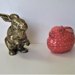 May include: A brass rabbit figurine stands next to a pink polka-dotted bunny-shaped trinket box. The brass rabbit is detailed with upright ears and a seated pose. The trinket box has a lid and is painted with white polka dots.