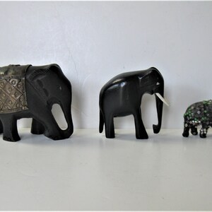 May include: Three decorative elephant figurines. The largest is black with a silver-colored saddle. The middle elephant is black with white tusks. The smallest elephant is black and covered in sequins and beads.