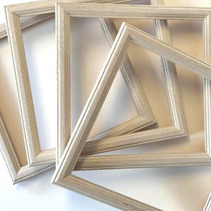 May include: A collection of four rectangular picture frames made of light-colored wood with a visible grain texture. The frames are arranged in an overlapping, layered composition, each featuring a simple, clean-lined molding design.