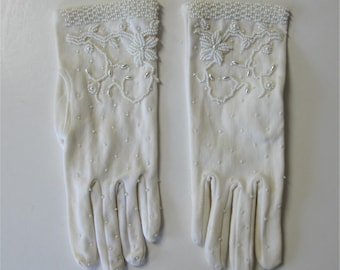 May include: A pair of ivory-colored gloves. The gloves are adorned with small, white beads along the cuffs and in a floral pattern. The gloves have a dotted texture and are likely made of a soft, breathable fabric. The gloves are designed for formal occasions.