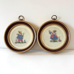 May include: Two circular wall hangings featuring embroidered figures in dark brown frames with gold-toned inner rims and brass hanging loops. The left piece shows a figure in a blue coat and pink pants with a staff, while the right piece shows a figure in a blue dress and pink apron holding a blue umbrella, both with small animals.