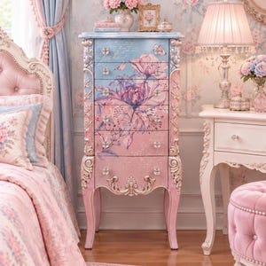 May include: A tall, slender wooden jewelry armoire featuring a gradient paint finish transitioning from light blue at the top to soft pink at the bottom. The front drawers are decorated with a large purple floral illustration, script text, and clear crystal knobs.