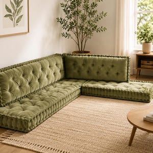 May include: An L-shaped floor sofa upholstered in olive green velvet fabric. The sofa features a tufted design with deep button detailing on both the seat cushions and the backrests, finished with thick, braided piping along the edges.