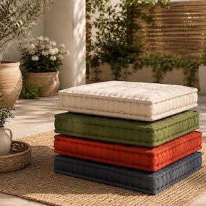 May include: A stack of four square floor cushions in cream, olive green, terracotta orange, and navy blue. Each cushion features a tufted design with visible stitching along the edges, creating a structured, boxy shape for floor seating.