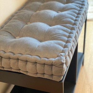 May include: A rectangular bench featuring a thick, tufted gray cushion with button detailing. The cushion has a piped edge and sits atop a black metal frame with a lower shelf, providing a structured and elevated seating surface for any room.