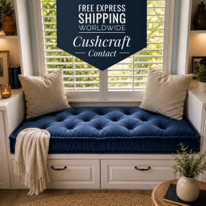 May include: A deep blue tufted bench cushion with a rectangular shape and button detailing, positioned on a white window seat. Two beige square throw pillows rest on the cushion, and a cream-colored fringed throw blanket hangs over the edge.
