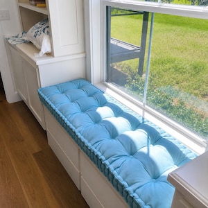 May include: A rectangular, light blue tufted bench cushion with a thick, piped edge. The cushion features a grid of fabric-covered buttons across its surface, creating a soft, dimpled texture. It is positioned on a white built-in window seat.