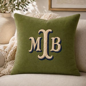 May include: A square olive green throw pillow featuring a central embroidered monogram with the letters M, I, and B. The letters are stitched in a light cream thread with a dark navy blue outline, creating a bold and textured decorative accent.