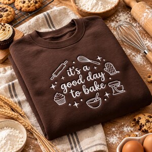 May include: A dark brown crewneck sweatshirt featuring white embroidered text that reads "it's a good day to bake" surrounded by small line-art icons of a rolling pin, whisk, oven mitt, stand mixer, cupcake, and mixing bowl.