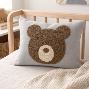 May include: A rectangular light grey throw pillow featuring a textured, tufted brown bear face design in the centre. The bear has rounded ears and a circular cream-coloured snout with a small brown nose detail, created with a punch needle technique.