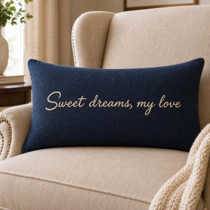 May include: A rectangular navy blue throw pillow featuring the embroidered text "Sweet dreams, my love" in a light cream-colored cursive script centered on the front surface. The pillow has a textured fabric finish and a simple rectangular shape.