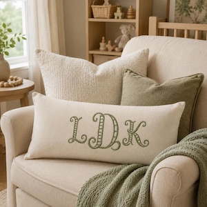 Puede incluir: Un cojín lumbar rectangular de color crema con un monograma bordado en verde oliva con las letras LDK en una fuente cursiva decorativa, colocado frente a un cojín cuadrado de textura crema y un cojín cuadrado de color verde oliva.