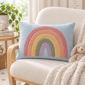 Puede incluir: Un cojín rectangular de color azul claro con un diseño de arcoíris con relieve en el centro. El arcoíris consta de arcos concéntricos en tonos suaves de rosa, naranja, amarillo, verde y morado, creados con un acabado de hilo texturizado.