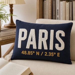 Pode incluir: Uma almofada retangular azul-marinho com a palavra PARIS em letras brancas grandes e texturizadas. Abaixo da palavra, as coordenadas geográficas 48,85° N / 2,35° E estão bordadas com fio amarelo dourado no centro da face da almofada.