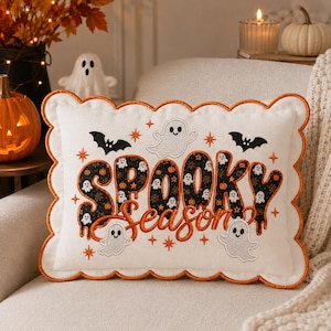 May include: A rectangular white throw pillow with a scalloped orange border. The front features embroidered text reading "SPOOKY Season" in black and orange, surrounded by stitched white ghosts, black bats, and small orange star accents.