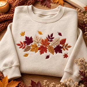 May include: A cream-colored crewneck sweatshirt featuring an embroidered design of various autumn leaves in shades of burgundy, orange, yellow, and tan centered on the chest. The long-sleeved garment has ribbed cuffs and a ribbed neckline.