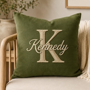 Può includere: Un cuscino quadrato verde oliva caratterizzato da una grande lettera K ricamata in beige come monogramma. Il nome Kennedy è cucito in un carattere beige corsivo al centro della lettera K. Il cuscino ha una consistenza del tessuto morbida e opaca.
