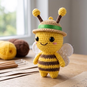 Puede incluir: Una figura de abeja tejida a ganchillo con cuerpo a rayas amarillas y marrones, alas blancas y ojos negros. La abeja lleva un sombrero color canela con una cinta verde y dos antenas con puntas amarillas. Tiene mejillas rosadas y una sonrisa.