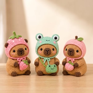 May include: Three crocheted capybara figures in tan yarn. The left one wears a pink strawberry hat and holds a strawberry. The center one wears a light green frog hat and a matching frog-shaped crossbody bag. The right one wears a pink peach hat.