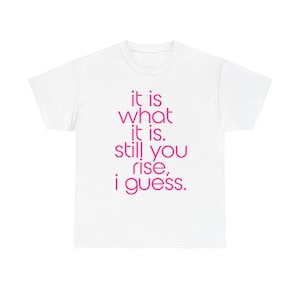 Può includere: Una t-shirt bianca a maniche corte con il testo "it is what it is. still you rise, i guess." stampato in un vivace carattere sans-serif rosa centrato sul petto. Il capo presenta un classico girocollo e maniche corte standard.