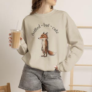 Könnte beinhalten: Ein beigefarbenes Sweatshirt mit Rundhalsausschnitt und einer grafischen Illustration eines Fuchses mit großen Augen sowie dem Text "Baffled - but - cute" in Schwarz über dem Tier. Das Sweatshirt hat lange Ärmel sowie gerippte Bündchen.