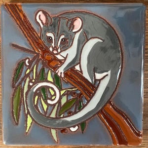 May include: A square ceramic tile featuring a glossy, raised-relief illustration of a grey and white possum perched on a brown tree branch. The possum has a long, curled tail and is surrounded by green eucalyptus leaves against a muted blue background.