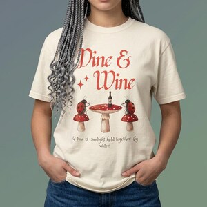 May include: A cream-colored t-shirt featuring the red text "Dine & Wine" above an illustration of two ladybugs sitting on mushrooms at a table with a wine bottle. Below the table, the text reads "Wine is sunlight held together by water."