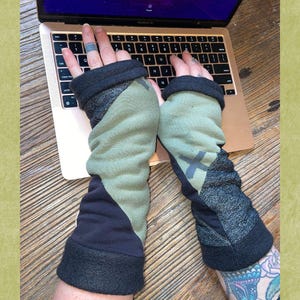 May include: A pair of fingerless arm warmers featuring a color-blocked design in olive green, charcoal gray, and black. The fabric has a soft, fleece-like texture with a dark gray cross graphic on the forearm. These sleeves extend from the knuckles to the mid-forearm, providing coverage while leaving the fingers exposed for typing.