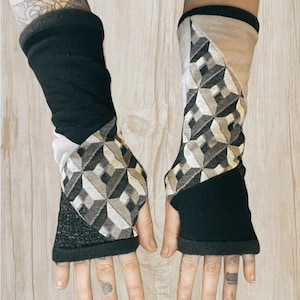 May include: A pair of fingerless gloves featuring a patchwork design of solid black fabric, a textured dark gray section, and a geometric pattern in shades of black, white, and beige. The gloves extend from the knuckles to the mid-forearm.