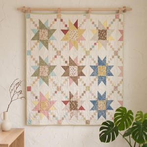 Könnte beinhalten: Ein rechteckiger Quilt mit einem sich wiederholenden Sternmuster aus verschiedenen floralen und geometrischen Stoffen in Gelb-, Rosa-, Blau-, Grün- und Brauntönen. Der Quilt hängt an einer Holzstange und hat einen cremefarbenen Hintergrund.