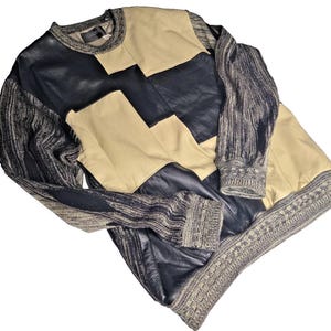 Puede incluir: Un suéter de manga larga con un diseño de bloques de color que presenta paneles de cuero negro y beige en la parte delantera y las mangas, combinado con un tejido de punto texturizado en gris jaspeado y negro en los costados, puños y cuello.