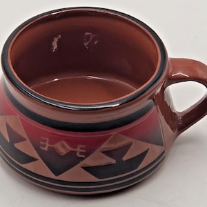 May include: A wide-mouthed ceramic mug with a glossy finish, featuring a terracotta base color and a decorative band of black and dark red geometric patterns. The mug has a single handle on the right side and a smooth, rounded interior surface.