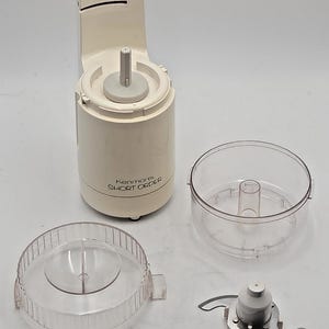 Peut inclure: Un robot culinaire Kenmore Short Order blanc avec un interrupteur marche-arrêt, un bol en plastique transparent, un couvercle en plastique transparent à bord strié et une lame en métal avec un moyeu central en plastique blanc pour la cuisine.