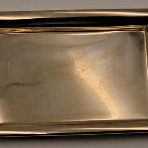 May include: A rectangular gold-toned metal tray with a polished finish and raised edges. The center of the tray features an engraved monogram with the letters RL and the text Ralph Lauren printed in a small, clear font below the initials.