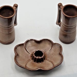 May include: A set of two tall, cylindrical brown ceramic mugs with vertical ribbed detailing and a matching brown ceramic candle holder shaped like a flower with a central circular opening for a candle, all featuring a smooth, glossy finish.