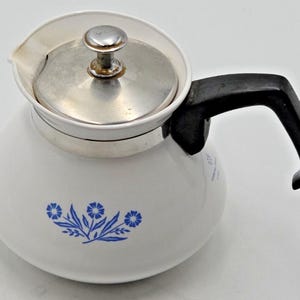 May include: A white teapot featuring a blue floral pattern on the front. The teapot has a silver-colored metal lid with a small knob handle and a black plastic handle attached to the side. The body is rounded with a wide base and a small spout.