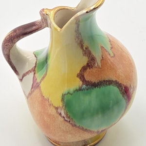 May include: A ceramic pitcher featuring a rounded body and a curved handle. The surface is decorated with a mottled, watercolor-style pattern in shades of yellow, green, and orange, separated by dark brown lines. The rim and base have gold trim.