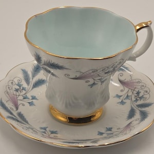 May include: A white porcelain teacup and saucer set featuring a scalloped edge with gold trim. The interior of the cup is light blue. The exterior and saucer are decorated with delicate blue and purple feathery floral patterns and gold accents.