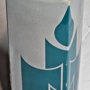 May include: A tall, rectangular cardboard box in a light gray color featuring a large, stylized teal blue graphic of a candle flame. The top label displays the text S3928 RASPBERRY / MULBERRY FRAMBOISE / MORE HIMBEERE / DUNKELPURPUR FRAMBUESA / MORA 2077.