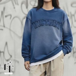 Puede incluir: Una sudadera azul de manga larga con cuello redondo y efecto de lavado desteñido. La palabra VICTORY está aplicada en el pecho con tela azul a juego y bordes deshilachados. Se ve una camiseta blanca en el dobladillo, combinada con pantalones beige.