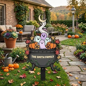 Puede incluir: Un estaca de jardín de metal negro con un caldero que lleva el texto THE SMITH FAMILY, lleno de tres fantasmas claros, calabazas naranjas y hojas de otoño. Una luna creciente se sitúa sobre los fantasmas y enredaderas rodean el caldero.
