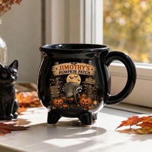 Puede incluir: Una taza de cerámica negra con forma de caldero que presenta un gráfico de un mapache en un huerto de calabazas con el texto "JIMOTHY'S PUMPKIN PATCH," "HAY RIDES," "CIDER," "CANDY & MORE!," y "PICK YOUR PERFECT PUMPKIN." Tiene tres patas.