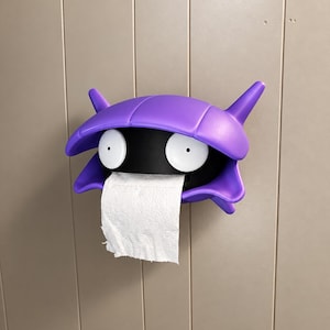 Puede incluir: Un portarrollos de papel higiénico de pared en color morado y negro con forma de criatura estilizada, con un caparazón rígido, dos orejas puntiagudas y dos grandes ojos circulares blancos. Un rollo de papel higiénico blanco cuelga del centro.