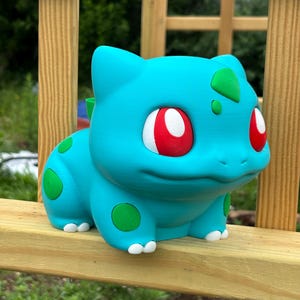 May include: A teal-coloured Bulbasaur figurine featuring a large, hollow opening on its back. The creature has bright red eyes, white claws, and distinct green circular spots on its body. The surface shows a subtle 3D-printed texture throughout.
