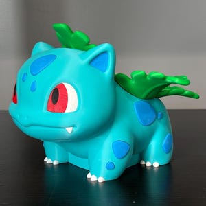May include: A teal-colored creature figurine with a large green plant bulb on its back. The figure features dark blue spots on its body, red eyes with black pupils, and small white fangs. It has four legs with white claws and a smooth finish.