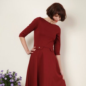 May include: A woman wearing a maroon dress with a boat neck and three-quarter sleeves. The dress has a fitted bodice and a flared skirt.