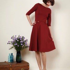 May include: A woman wearing a maroon dress with a fitted bodice and a full skirt. She is standing on a wooden floor and has a bouquet of purple flowers in a vase on a wooden chest behind her.